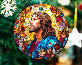 May include: Round Christmas ornament featuring a stained-glass design of a man with long brown hair and a beard, wearing a blue robe. The ornament is decorated with colorful flowers and set against a yellow and orange background.