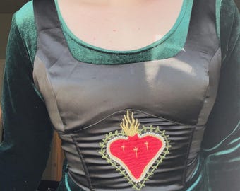 May include: A dark green velvet and black satin bodice top with long sleeves. The bodice features an embroidered red heart with gold flames and three crosses. The neckline is trimmed in green velvet.