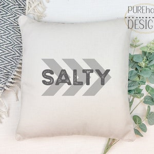 Könnte beinhalten: Ein weißes Kissen mit dem Wort "SALTY" in grauem Druck mit einem Used-Look. Das Wort ist mittig auf dem Kissen platziert und von drei grauen Pfeilen umgeben, die nach rechts zeigen.