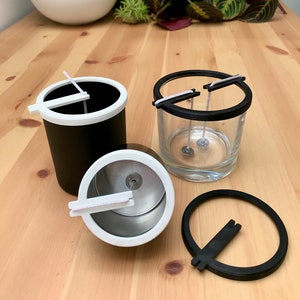 Peut inclure: Ensemble de fournitures pour la fabrication de bougies, comprenant des récipients noirs et blancs, un verre transparent et des supports de bougies noirs et blancs. Les articles sont disposés sur une surface en bois, avec des plantes en arrière-plan.