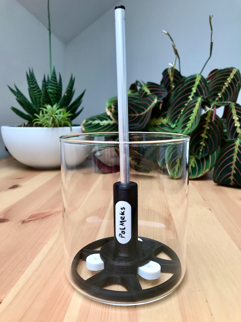 May include: A clear glass beaker with a black and white device inside. The device has a black base with white circular components and a silver rod extending upwards. The text "PolMeks" is visible on a white label. Plants are in the background.