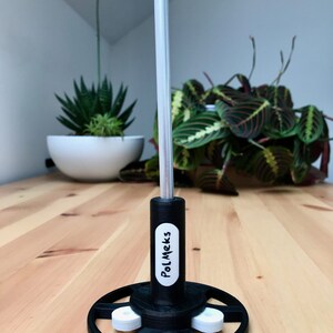 May include: A black and white stand with the text "PolMeks" on the side. The stand has a circular base with three white supports and a tall, silver pole extending upwards. The stand is on a wooden surface.