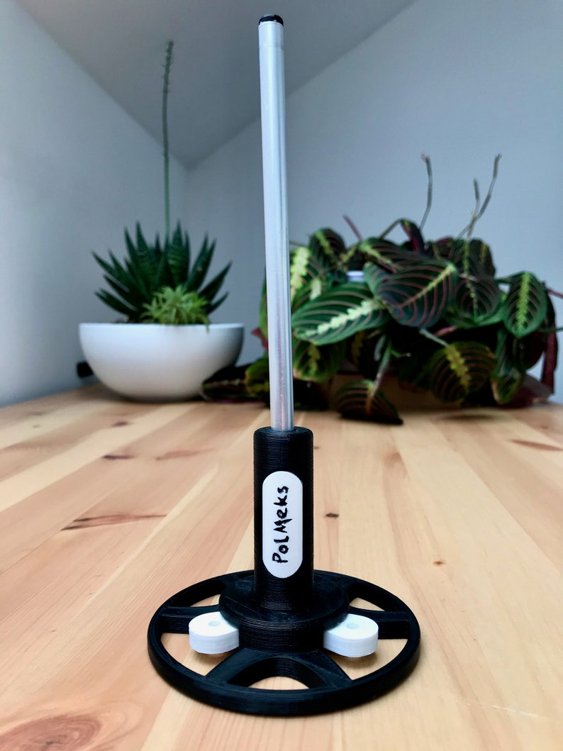 May include: A black and white stand with the text "PolMeks" on the side. The stand has a circular base with three white supports and a tall, silver pole extending upwards. The stand is on a wooden surface.