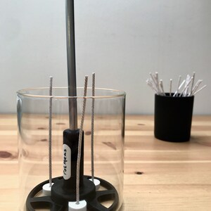 May include: A clear glass candle holder with a black base and a metal rod in the center. The holder contains several wicks and is next to a small black container filled with additional wicks. The text "POLMEKS" is visible on the holder.