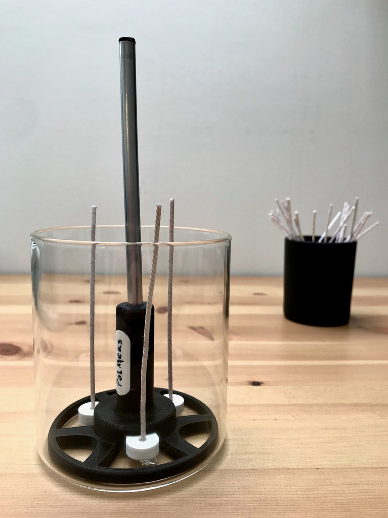 May include: A clear glass candle holder with a black base and a metal rod in the center. The holder contains several wicks and is next to a small black container filled with additional wicks. The text "POLMEKS" is visible on the holder.