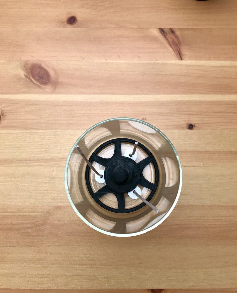 May include: A clear glass candle holder with a black, star-shaped insert and three wicks. The candle holder sits on a light brown wooden surface. The wicks are a light brown color.