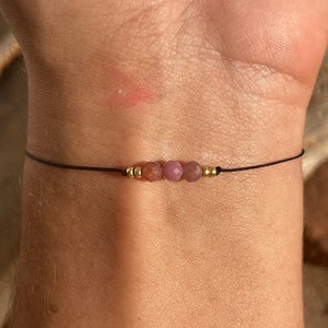 May include: A simple bracelet with a black cord and four small pink and gold beads.