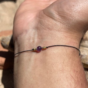 May include: A simple bracelet with a single purple gemstone bead and gold spacer beads on a black cord.
