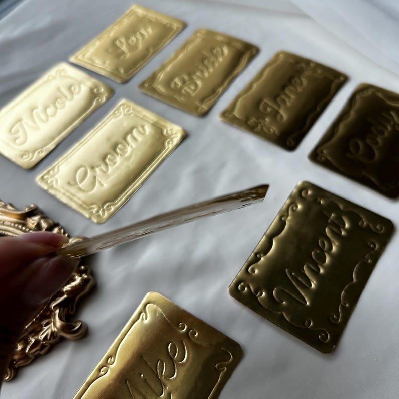 May include: A collection of rectangular gold-colored name plates featuring embossed script text including Nicole, Groom, Bride, Jane, Cody, Vincent, and Aimee. Each plate has a decorative scalloped border. One plate is held up to show its thin profile.
