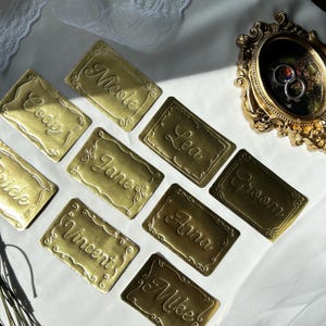 May include: A collection of rectangular gold-colored metal place cards featuring embossed names including Cody, Nicole, Lea, Jane, Groom, Bride, Vincent, Anna, and Mike, accompanied by an ornate gold oval frame containing two silver rings.