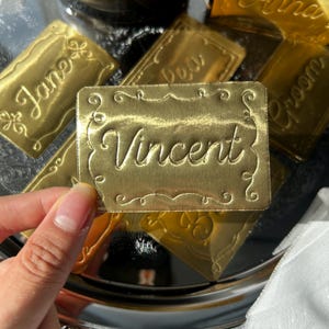 May include: A rectangular gold-colored metal name tag featuring the name Vincent engraved in a cursive script font. The tag has a decorative scalloped border with ornate scrollwork details at the corners and edges, creating a polished finish.