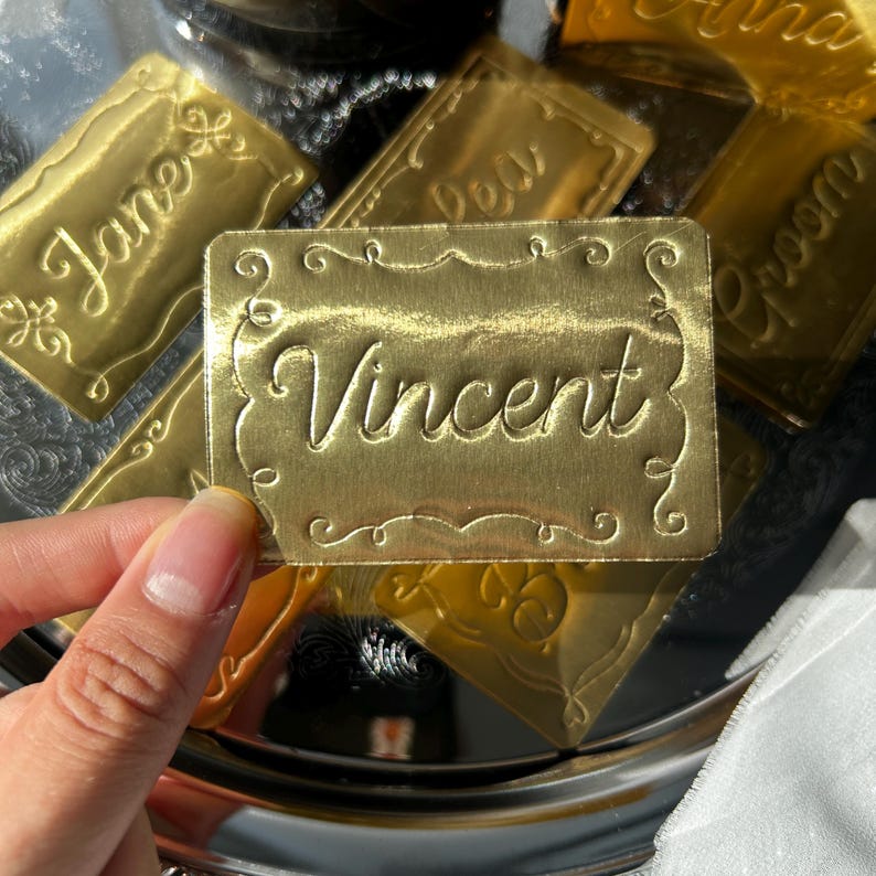 May include: A rectangular gold-colored metal name tag featuring the name Vincent engraved in a cursive script font. The tag has a decorative scalloped border with ornate scrollwork details at the corners and edges, creating a polished finish.