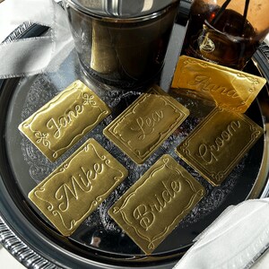 May include: A collection of rectangular gold-colored place cards with ornate embossed borders and cursive lettering. The cards feature the names Jane, Lea, Mike, Bride, Groom, and Anna, arranged on a reflective silver-toned circular tray.