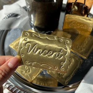 May include: A rectangular gold-colored metallic tag with a decorative embossed border and the name Vincent written in a cursive script. The tag has a reflective surface and is held up to show the detailed engraving and the metallic finish.