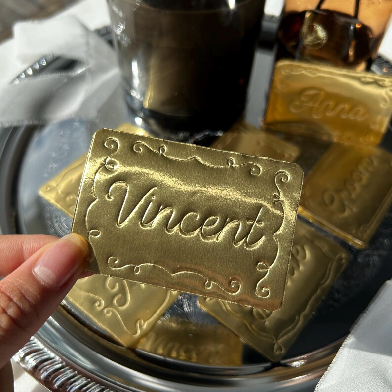 May include: A rectangular gold-colored metallic tag with a decorative embossed border and the name Vincent written in a cursive script. The tag has a reflective surface and is held up to show the detailed engraving and the metallic finish.