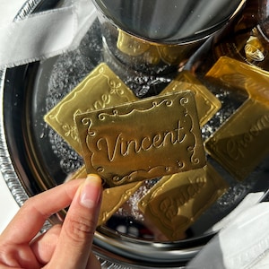 May include: A rectangular gold-colored metal tag with a decorative embossed border and the name Vincent engraved in a cursive script. The tag is held against a background of a silver tray containing several other similar gold-colored metal tags.