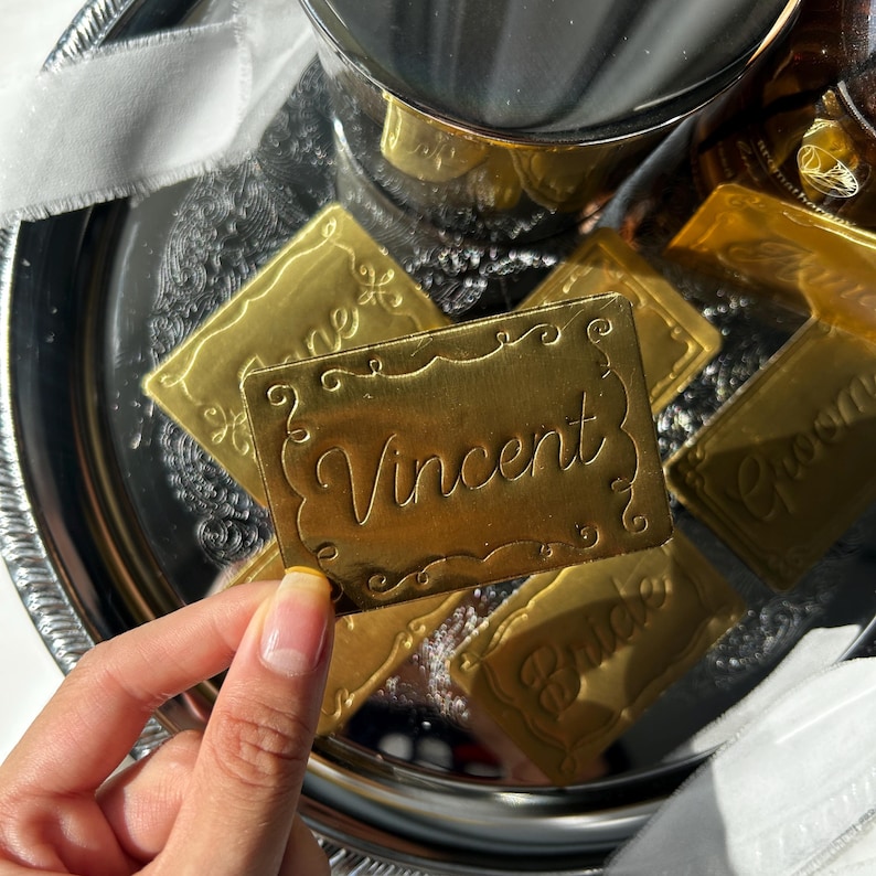 May include: A rectangular gold-colored metal tag with a decorative embossed border and the name Vincent engraved in a cursive script. The tag is held against a background of a silver tray containing several other similar gold-colored metal tags.