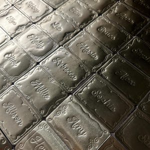 May include: A collection of rectangular gold-toned metal tags arranged in a grid. Each tag features an embossed decorative border and a unique name engraved in a cursive script, including names such as Jordan, Mike, Kelsey, Kim, Molly, and Amy.