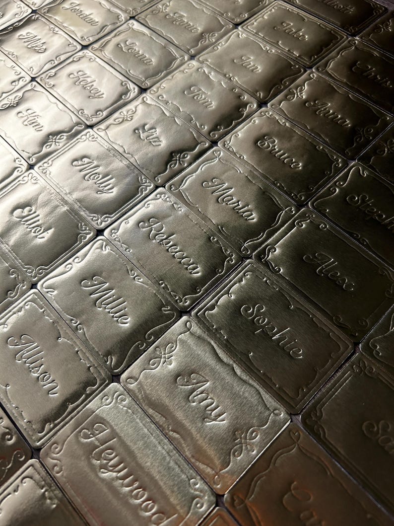 May include: A collection of rectangular gold-toned metal tags arranged in a grid. Each tag features an embossed decorative border and a unique name engraved in a cursive script, including names such as Jordan, Mike, Kelsey, Kim, Molly, and Amy.