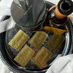 May include: Five rectangular brass place cards with scalloped edges and embossed cursive names including Jane, Lea, Groom, Mike, and Bride. These metallic tags are arranged on a dark, reflective circular tray alongside a Zara Home candle.