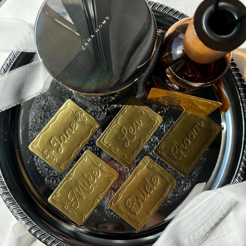 May include: Five rectangular brass place cards with scalloped edges and embossed cursive names including Jane, Lea, Groom, Mike, and Bride. These metallic tags are arranged on a dark, reflective circular tray alongside a Zara Home candle.