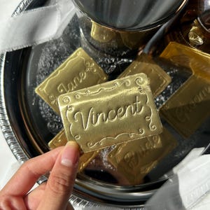 May include: A rectangular gold-colored place card with a decorative embossed border and the name Vincent written in a cursive script. The card is one of several similar gold place cards featuring various names and titles in a matching style.