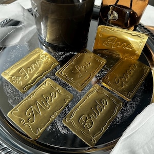 May include: A collection of rectangular gold-colored metal place cards featuring ornate embossed borders and engraved names including Anna, Jane, Lea, Mike, Groom, and Bride in a cursive script font, arranged on a reflective silver-toned circular tray.