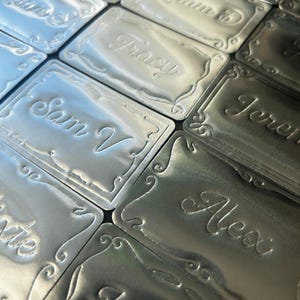 May include: A collection of rectangular silver-toned metal tags featuring embossed decorative borders and various names engraved in a cursive script, including Sam V, Tracy, Adam B, Paul, Jeremy, and Alex, arranged in a grid-like pattern.