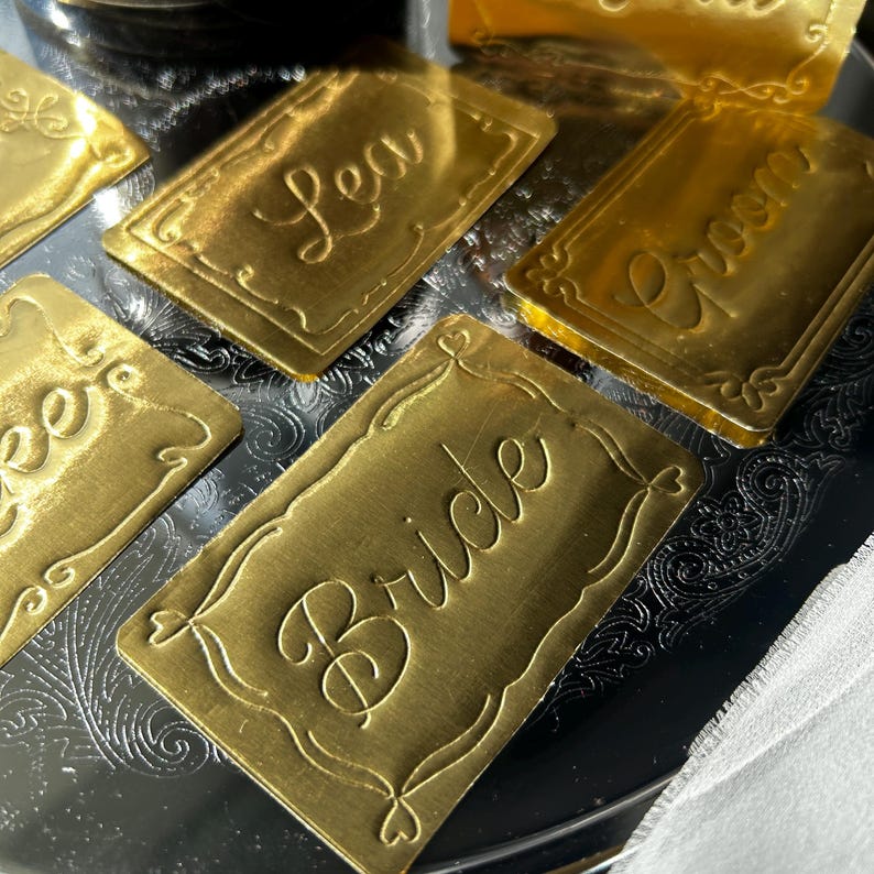 May include: A collection of rectangular gold-colored metal place cards featuring embossed script text. The visible cards display the words Bride, Groom, and Lea, each framed by a decorative border with scrollwork details at the corners.