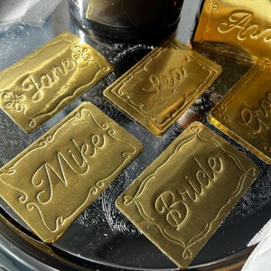 May include: A collection of rectangular gold-colored name plates featuring embossed script lettering. The plates display the names Jane, Lea, Mike, and Bride, each framed by a decorative border with small flourishes and heart-shaped accents.