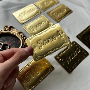 May include: A collection of rectangular gold-colored metallic name tags with embossed borders and cursive text including Nicole, Bride, Groom, Jane, Mike, and Vincent. Each tag features decorative flourishes at the corners and edges.