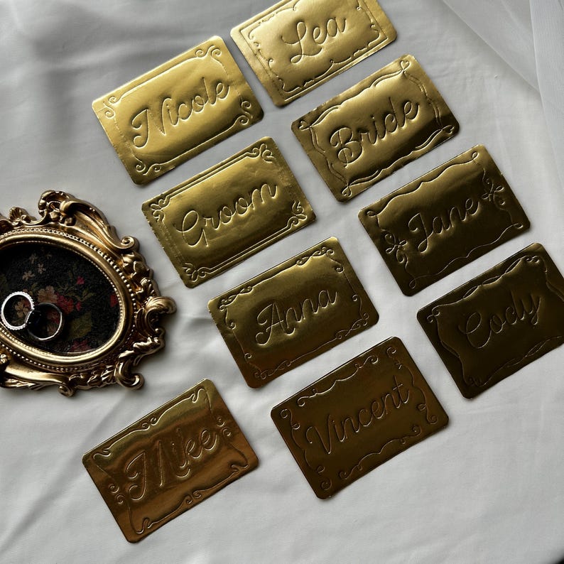 May include: A collection of rectangular gold-colored metal name plates with embossed script text including Nicole, Lea, Bride, Groom, Jane, Anna, Cody, Mike, and Vincent, alongside an ornate oval gold frame containing two silver rings.