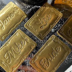 May include: A collection of rectangular gold-toned metal name tags featuring embossed script lettering. Each tag displays a unique name or title, including Jane, Lea, Mike, and Bride, surrounded by a decorative embossed border frame.