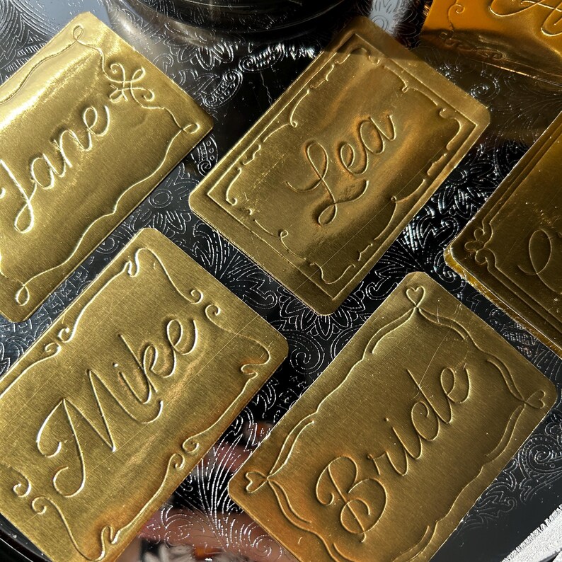 May include: A collection of rectangular gold-toned metal name tags featuring embossed script lettering. Each tag displays a unique name or title, including Jane, Lea, Mike, and Bride, surrounded by a decorative embossed border frame.
