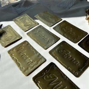 May include: A collection of rectangular brass-colored metal place cards featuring embossed decorative scalloped borders and engraved names including Cody, Nick, Lea, Bride, Jane, Vincent, Anna, and Mia, arranged on a white surface.