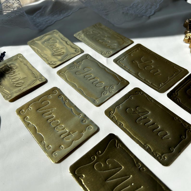 May include: A collection of rectangular brass-colored metal place cards featuring embossed decorative scalloped borders and engraved names including Cody, Nick, Lea, Bride, Jane, Vincent, Anna, and Mia, arranged on a white surface.
