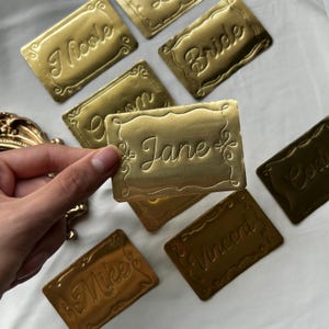 May include: A collection of rectangular gold-colored metallic name tags with embossed decorative borders. Each tag features a unique name in cursive script, including Nicole, Bride, Jane, Mike, Vincent, and Cody, with one tag held in the foreground.