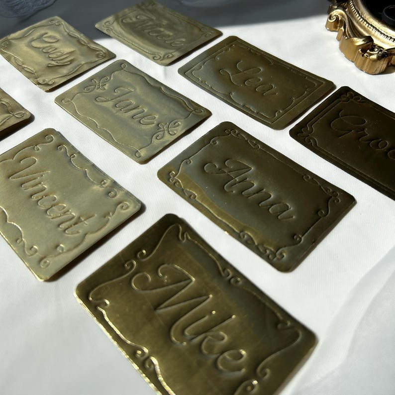 May include: A collection of rectangular brass-colored metal place cards, each featuring an embossed decorative border and a unique name in cursive script, including names such as Cody, Jane, Vincent, Lea, Anna, and Mike, arranged on a white surface.