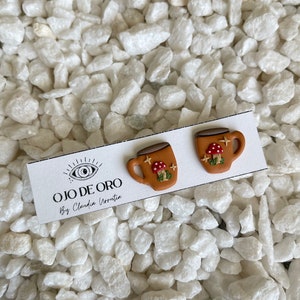 May include: A pair of brown ceramic earrings shaped like mugs with white mushrooms and a white star design. The earrings are on a white card with the text "OJO DE ORO By Claudia Ivontia".