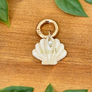 May include: A gold-tone seashell charm with a white pearl accent and the word "Pearl" inscribed on it. The charm is attached to a gold-tone lobster clasp.