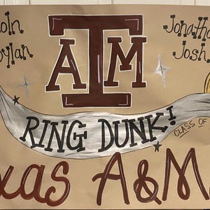 May include: A brown poster with the text "Texas A&M University" and "Ring Dunk!" in a stylized font. The poster includes the letters "A&M" and the names "Lincoln" and "Dylan". A ring is depicted with the text "Class of 2025".