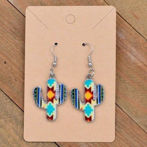 Peut inclure: Une paire de boucles d'oreilles pendantes argentées avec un motif de cactus. Le cactus est jaune avec des motifs géométriques bleus, rouges et orange.