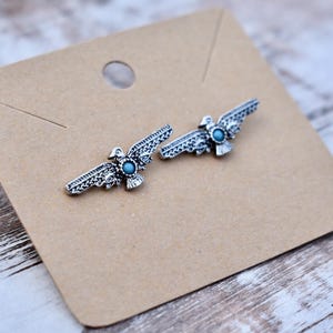 May include: A pair of silver-toned eagle-shaped earrings with turquoise-colored stone accents, displayed on a brown card. The earrings have detailed wing and body designs. The card is on a light-colored wooden surface.