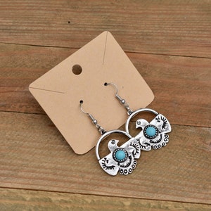 May include: Silver dangle earrings with turquoise stones and a decorative design. The earrings are on a brown paper card.