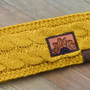May include: A yellow knitted headband with a brown leather patch featuring a sunflower design. The patch also has the letters "C.C." on it.