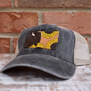 May include: A black and brown trucker hat with a patch featuring a brown buffalo and yellow sunflowers. The patch is on a tan background.