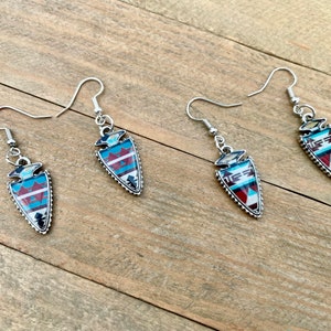 Peut inclure: Quatre boucles d'oreilles argentées avec un motif de pointe de flèche. Chaque boucle d'oreille présente un motif géométrique turquoise, rouge et blanc dans un cadre argenté. Les boucles d'oreilles ont une fermeture à crochet.