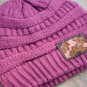 May include: A mauve knitted beanie with a brown leather patch featuring a floral design. The patch has the letters "CC" on it.