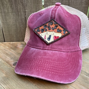 May include: A burgundy and beige trucker hat with a leather patch featuring three playing cards, an ace of spades, an ace of diamonds, and an ace of hearts. The patch is diamond-shaped and has a brown leather background with a leopard print pattern.