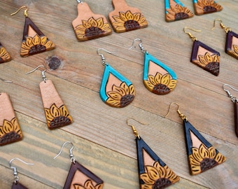 May include: A collection of handmade leather earrings featuring sunflower designs. The earrings are available in a variety of shapes and colors, including brown, black, teal, and tan.